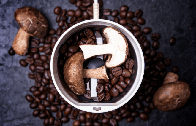 Top view of coffee beans and mushrooms in a coffee grinder with more coffee beans and mushrooms on the counter surrounding the grinder. 